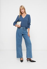 Blå langermet V-hals topp kombinert med høytlivede, vide lyseblå jeans. Tilbehør inkluderer en hvit klut og svarte spisse hæler.