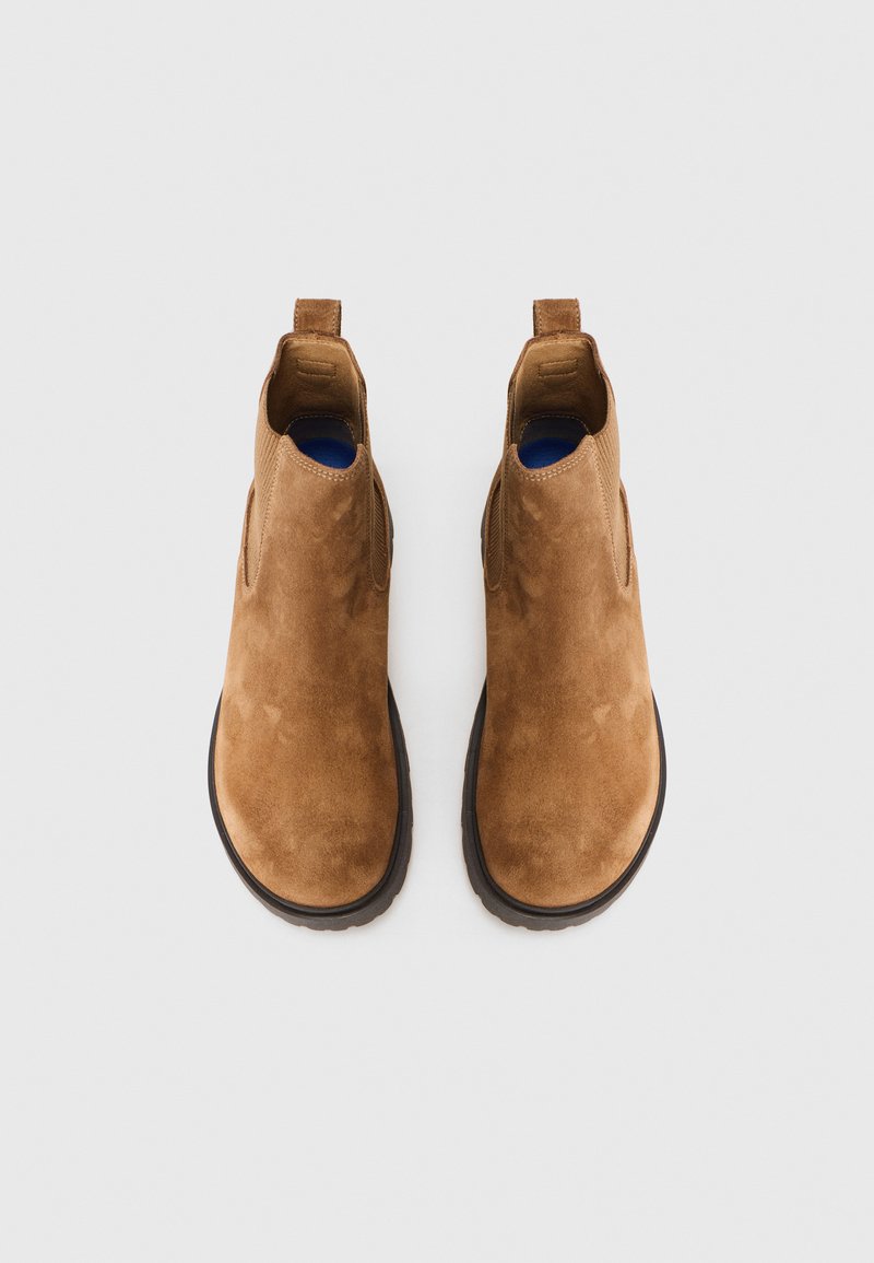 Una coppia di stivali Chelsea in suede marrone con suole nere, visti dall'alto su uno sfondo bianco uniforme.