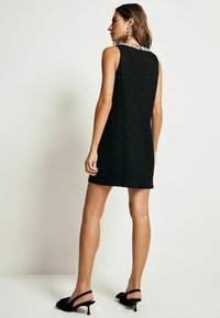 Robe noire sans manches en tissu texturé, avec un décolleté orné de perles et une coupe droite, associée à des talons noirs à lanières.