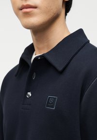 Κοντινό πλάνο ατόμου που φοράει μπλε ναυτικό πόλο με γιακά, δύο κουμπιά και μικρό κεντημένο λογότυπο στο αριστερό στήθος.