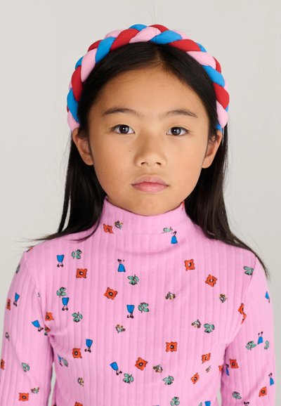 Jeune fille aux cheveux noirs et raides portant un t-shirt rose à col montant avec de petits imprimés floraux et un bandeau tressé rouge, bleu et rose.
