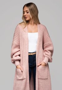 Dlouhý, volně pletený cardigan v jemně růžové barvě s oversized rukávy, texturovanou tkaninou a předními kapsami, nošený přes bílý top a tmavé džíny.