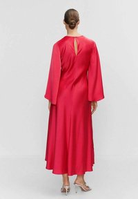 Robe longue en satin rouge avec des manches évasées et un dos en trou de clé, ourlet fluide et texture lisse. Portée avec des sandales à talons argentées.