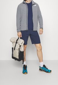 Grå fleecetröja, marinblå skjorta, blå shorts och turkosa träningsskor. Beige och svart rulltoppsryggsäck med justerbara remmar.