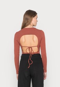 Camisola de manga longa em tom rust, com uma grande abertura nas costas, detalhe de laço ajustável e textura canalizada. Combinada com calças pretas de cintura alta.