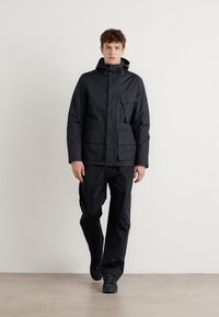 Veste noire imperméable avec capuche et multiples poches, associée à un pantalon et des chaussures noires assortis, présentée sur un fond uni.