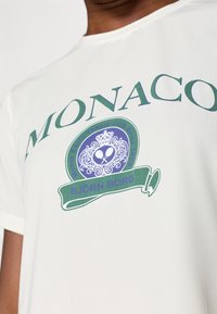 Fehér, rövid ujjú póló, amelyen egy nagy zöld és lila grafika található a "MONACO" felirattal és egy koronával, valamint az alján a "BJÖRN BORG" felirattal.