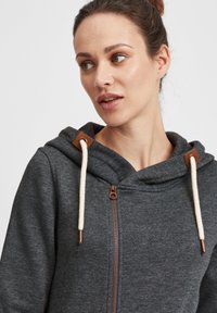 Grå zip-up hoodie med en mjuk textur, krämfärgade snoddar och bruna metallaccenter vid huvan, med en avslappnad passform och modern design.