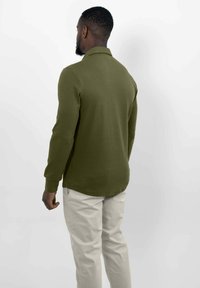 Een man die met zijn rug naar de camera staat, gekleed in een olijfgroen long-sleeve shirt en lichtbeige broek, tegen een effen witte achtergrond.