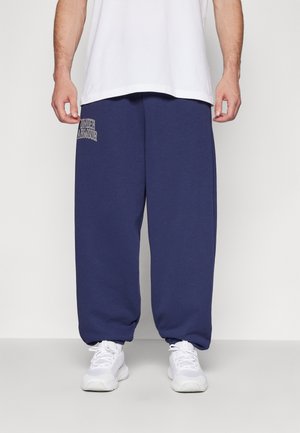 Man wearing navy blue Under Armour sweatpants, white sneakers, and a white t-shirt, standing against a plain background.