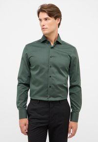 Camicia da uomo in tessuto verde scuro con colletto button-down, bottoni neri e maniche lunghe, abbinata a pantaloni neri.