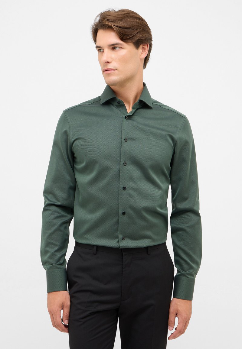 Camicia da uomo in tessuto verde scuro con colletto button-down, bottoni neri e maniche lunghe, abbinata a pantaloni neri.