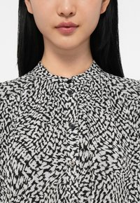 Γυναίκα με ίσια μαύρα μαλλιά, ντυμένη με μπλούζα με ψηλό λαιμό, που έχει μαύρη και άσπρη αφηρημένη εκτύπωση με κηλίδες και μαύρα κουμπιά.