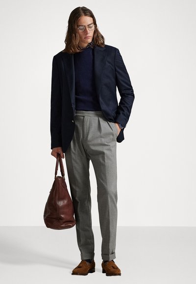 Blazer azul-marinho sobre um suéter azul-marinho, calças cinza-claro, bolsa de couro castanho e sapatos de camurça castanhos. O modelo tem cabelo comprido e usa óculos.