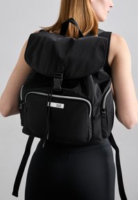 Black nylon backpack with a fold-over flap, front zip pocket, metal hardware, and adjustable padded shoulder straps. Features a logo tag.