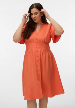 Kvinde justerer hår iført knælængde orange kjole med knapper foran, korte ærmer og figursyet talje mod hvid baggrund.