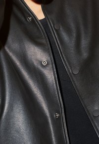 Veste en cuir noir à texture lisse, boutons pression et col classique, portée sur une chemise noire.