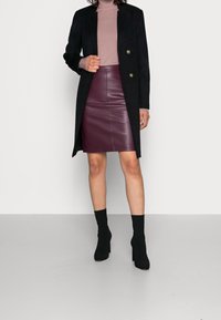 Svart lång kappa med guldknappar, ljuslila polotröja och en burgunderfärgad läderpennkjol. Svarta ankelstövlar fulländar outfiten.