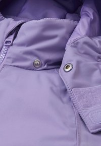 Jasnofioletowa kurtka z gładką, wodoodporną powierzchnią, odsłoniętym zamkiem błyskawicznym, napami oraz detalem z rzepem na kołnierzu.