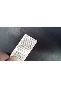 Main tenant une étiquette d’entretien de tissu indiquant 100 % coton et les instructions de lavage en plusieurs langues sur une surface sombre texturée.