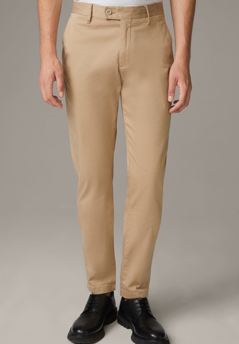 Beige Chinos aus leichtem Stoff, mit schlanker Passform, glatter Textur und vorderem Knopfverschluss. Kombiniert mit schwarzen Schuhen.