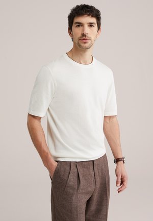 Homme aux cheveux courts et bouclés portant un pull blanc à manches courtes et un pantalon plissé marron, debout avec une main dans la poche.