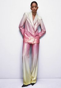 Donna che indossa un completo su misura lucido con ombre da rosa a giallo, composto da pantaloni a gamba larga e un blazer doppiopetto, in piedi contro un muro bianco.