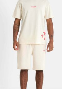 Beige cotton t-shirt and shorts set featuring a small pink logo on the chest and pink abstract patterns on the shorts.