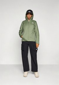 Zelena jakna z zadrgo in kapuco ter sprednjimi žepi, kombinirana s črnimi cargo hlačami z več žepi. Lahki materiali.