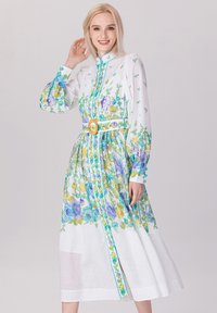 Robe à fleurs avec une base blanche, ornée de fleurs bleues et vertes. Manches longues, fermeture à boutons à l'avant, taille cintrée et large ceinture jaune.