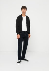 Svart strikket cardigan med knappelukking, hvit t-skjorte, mørke denimjeans og svarte sneakers. Modellen står foran en nøytral bakgrunn.
