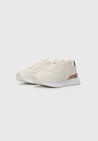 Paire de baskets couleur crème avec semelles blanches, design à lacets et accent métallisé bronze sur le talon, sur fond blanc uni.