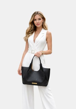 Donna con lunghi capelli biondi mossi indossa una tuta bianca senza maniche con cintura, gioielli d'oro e tiene una grande borsa nera con manici intrecciati.