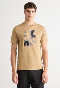 Beige bomull t-skjorte med et blokk-mønster grafikk i svart og hvitt på fronten, rund halslinning og korte ermer.