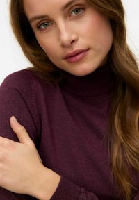 Vihreäsilmäinen nainen, jolla on pitkät ruskeat hiukset ja violetti poolopaita, pitäen vasenta kättään hellästi.