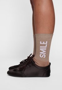 Chaussures marron à lacets associées à des chaussettes pailletées beiges arborant le mot "SMILE" en lettres blanches. Accent sur la texture et la visibilité des jambes.