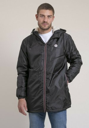 Homme portant un parka noir à capuche avec une doublure en polaire blanche, zippé sur une chemise blanche, debout avec une main dans la poche.