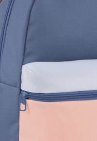 Zaino con tessuto blu, rosa e grigio chiaro. Presenta una tasca con zip sulla parte frontale, realizzata in materiale testurizzato e resistente.
