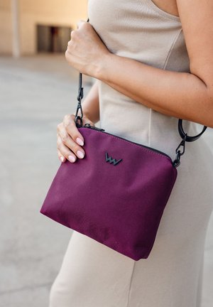 Lila Stofftasche mit einem schwarzen Reißverschluss und Logo. Verfügt über einen abnehmbaren schwarzen Riemen, eine glatte Textur und eine rechteckige Form.