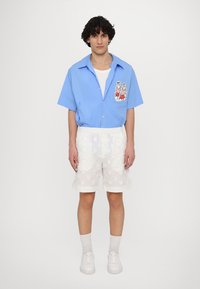 Blå kortærmet skjorte med knaplukning og grafisk lomme-design; hvide strukturerede shorts med indviklede mønstre; båret med hvide sneakers.