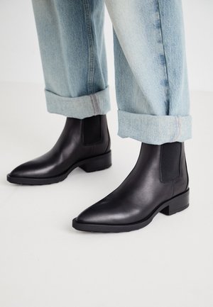 Zwarte leren enkellaarsjes met spitse neuzen en elastische zijpanelen, gecombineerd met lichtblauwe opgerolde denim jeans, wat samen een casual outfit vormt.