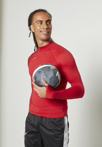 Rotes Langarm-Aktionsshirt mit tailliertem Schnitt, kombiniert mit schwarzen Shorts mit weißen Akzenten, während ein schwarzer Fußball gehalten wird.