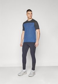 Blue and black athletic t-shirt with short sleeves, textured fabric, and logo on the chest, paired with dark leggings and light sneakers.