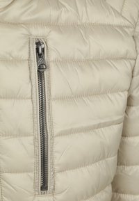 Beige pufferjack met horizontaal gewatteerde textuur, voorzien van een centrale rits in donkere metalen hardware voor sluiting. Glad, glanzend stoffen afwerking.