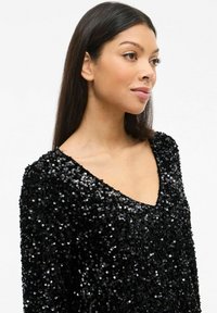 Juodas blizgančiais siūlais siuvinėtas viršus su V formos iškirpte, turi prigludusią siluetą ir tekstūruotą paviršių iš blizgančių apvalių blizgučių.