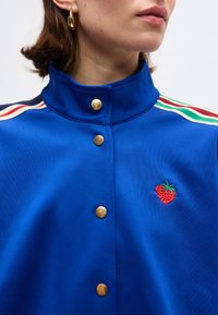 Κοντινό πλάνο ενός ατόμου που φορά μπλε σακάκι με χρυσά κουμπιά, πολύχρωμες ρίγες στους ώμους και μια μικρή κεντητή φράουλα στο στήθος.