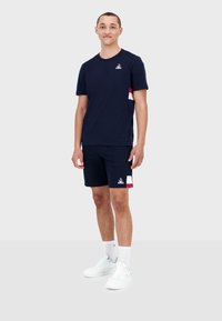 Ensemble t-shirt en coton marine et shorts, avec un logo sur la poitrine et des poches latérales avec des accents rouges et blancs. Baskets blanches et chaussettes.