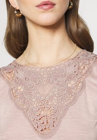 Ljusrosa topp med en dekorativ spetslivstycke som har intrikata blommönster och en texturerad finish. Guldhalsband och hoopsringar.