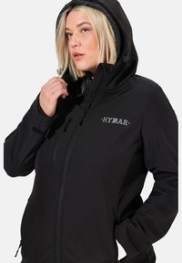 Giacca softshell nera con cappuccio, dotata di zip frontale, tasche laterali e un interno strutturato. Logo "HYRAR" sul petto.