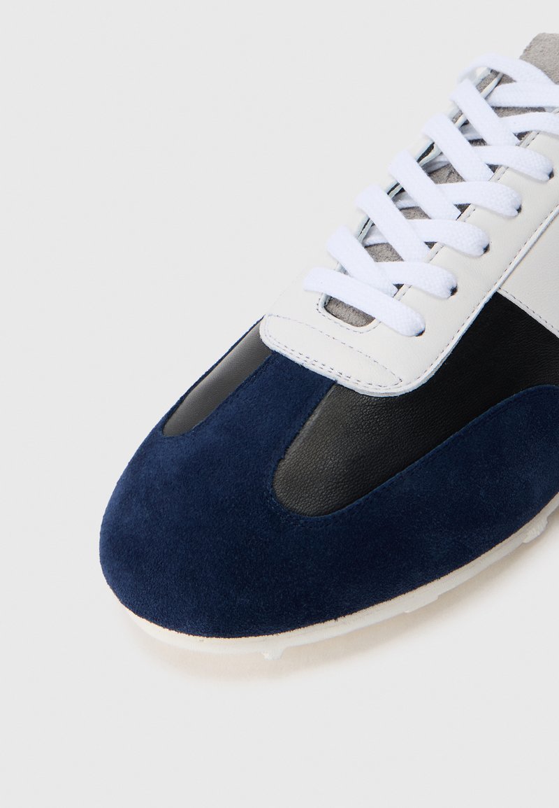 Sneaker med navy ruskind overdel, sorte læderdetaljer, hvide snørebånd og grå ruskind tunge. Rund tå og tekstureret såledetalje synlig.